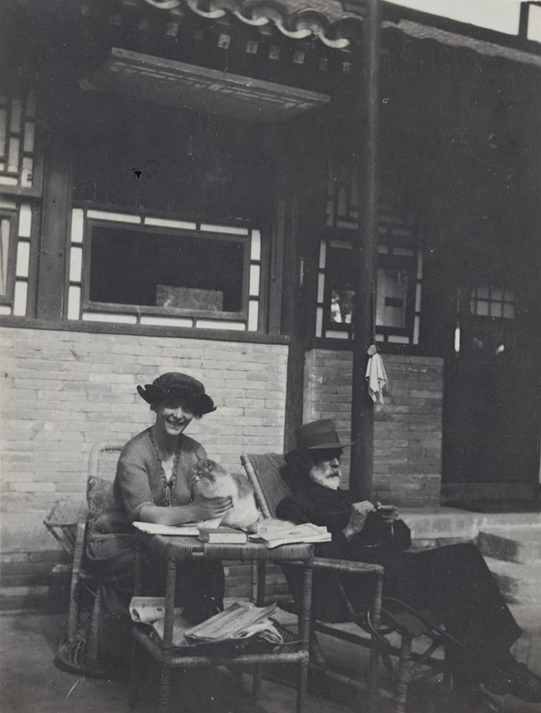 Guy, knitting at Balizhuang, late autumn 1921. Books are on the table and Ella is holding their cat Carpentier. HPC ref: EH01-343.